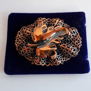 May include: A copper and silver toned brooch featuring a stylised leaf design. The leaf is set in a delicate filigree frame.
