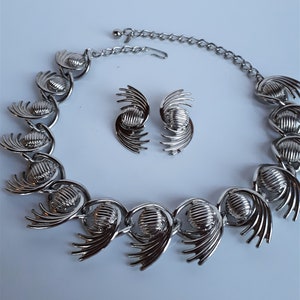 Può includere: Un set di collana e orecchini in tono argento con un design geometrico. La collana è realizzata con cerchi collegati con un design a ventaglio su ogni lato. Gli orecchini hanno un design abbinato.