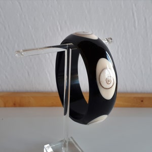 May include: Black bangle bracelet with white shell inlays. The bracelet is on a clear display stand.