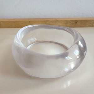 Könnte beinhalten: Ein klarer, runder Armreif. Das transparente Acrylmaterial reflektiert das Licht und erzeugt Glanzlichter und Schatten. Das Armband hat eine glatte, polierte Oberfläche und ein dickes, substanzielles Design. Dieses Accessoire ist für den Alltag geeignet.