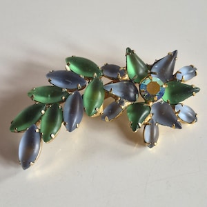 May include: A floral-shaped brooch featuring marquise-cut frosted glass stones in shades of green and periwinkle blue, set in a gold-tone metal frame. A single iridescent, faceted round crystal is positioned at the centre of the flower cluster.