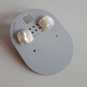 May include: A pair of white, square-shaped earrings on a grey card with the text "click-six.com" and "six" printed on it.