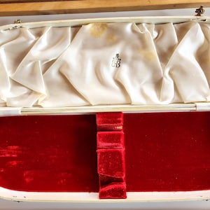 May include: An open white box with a red velvet interior. The box has a gold latch and a gold inscription on the top.