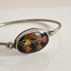 Peut inclure: Bracelet jonc argenté avec un élément central ovale. L'élément central présente des fleurs orange séchées sur fond noir. Le bracelet a un design simple et élégant, adapté au quotidien.