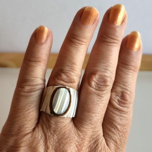 May include: A silver ring with an oval mother-of-pearl inlay with a striped design. The ring is worn on a hand with orange nail polish. The ring has a rectangular band with a textured surface.