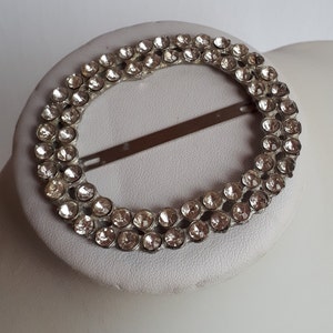 May include: A silver oval-shaped brooch with a double row of clear rhinestones.