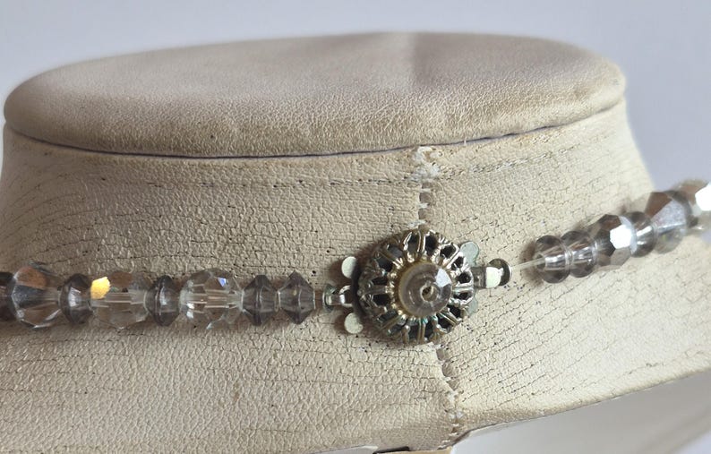 K&ouml;nnte beinhalten: Eine Halskette mit klaren, facettierten Kristallperlen, die auf einer d&uuml;nnen, transparenten Schnur aufgereiht sind. Die Kette verf&uuml;gt &uuml;ber einen runden, silberfarbenen Metallverschluss mit filigranem Durchbruchmuster und einer kleinen Perle.