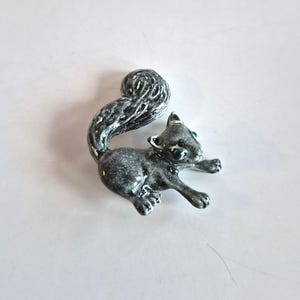 May include: A gray squirrel brooch with a curled tail and green eyes. The squirrel is in a running pose, with its front paws extended. The brooch is made of a shiny material.