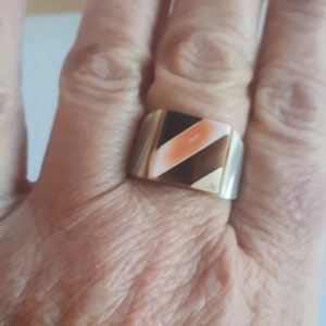 Peut inclure: Une bague de forme carrée avec une pierre multicolore sertie dans une bande dorée. La pierre est un mélange de blanc, de brun et d'orange.