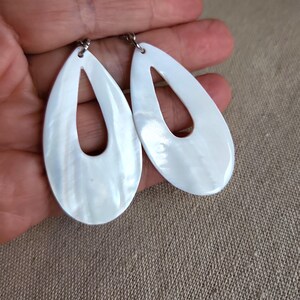 May include: A pair of white oval-shaped earrings with a cutout design. The earrings are made of mother of pearl and have a silver tone metal hook.