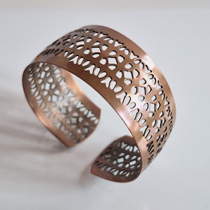 Könnte beinhalten: Ein breites Kupferarmband mit offenem Design. Das Armband hat eine gebogene Form und weist ein sich wiederholendes Muster aus organischen Formen auf. Das Metall hat eine warme, rötlich-braune Farbe und eine polierte Oberfläche.