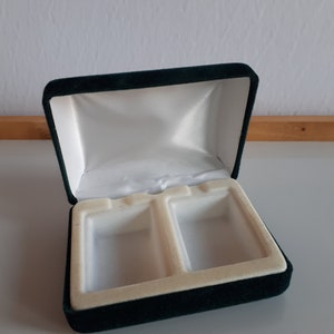 Puede incluir: Una caja de terciopelo verde oscuro con dos compartimentos, forrados con terciopelo blanco. La caja está abierta y vacía.