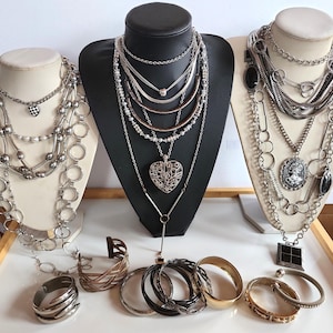 Può includere: Una collezione di collane e braccialetti esposti su busti di gioielli bianchi e neri. Le collane presentano vari design, tra cui catene, perline e ciondoli. Sono visibili anche braccialetti in argento, oro e nero.