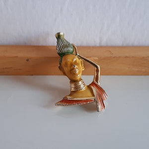 Puede incluir: Broche vintage que representa a una mujer que lleva un recipiente de agua. El broche tiene un acabado de metal dorado con detalles de esmalte en amarillo, naranja y verde. La mujer lleva un collar dorado. El broche es probablemente de mediados del siglo XX.
