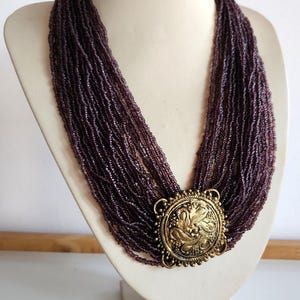 May include: A multi-strand necklace with small, dark purple beads. The necklace features a large, ornate, gold-coloured pendant with a floral design. The necklace is displayed on a white mannequin bust.