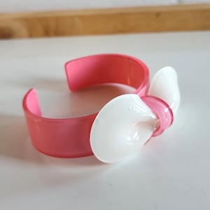 May include: A pink plastic bracelet with a white bow. The bracelet is made of a smooth, shiny plastic. The bow is attached to the bracelet with a small metal pin.