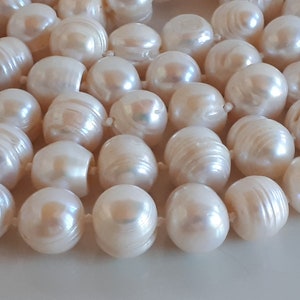 May include: Close-up of a strand of lustrous, off-white pearls. The pearls are irregularly shaped with a smooth, iridescent surface. The pearls are strung together, creating a necklace or bracelet.