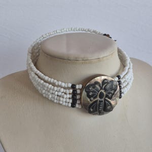 Könnte beinhalten: Weißes Choker-Halskette mit einem großen, runden Anhänger mit einem geschnitzten Schmetterlingsdesign. Der Anhänger ist eine Mischung aus Schwarz und schillernden Farben. Die Halskette hat mehrere Stränge aus weißen Perlen mit schwarzen Akzentperlen.