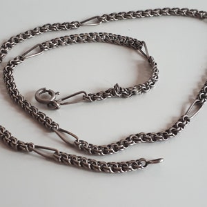 May include: A silver necklace with a fine chain design. The necklace has small, interconnected links, with elongated, oval-shaped links. A clasp is visible at one end. The necklace is displayed on a white surface.