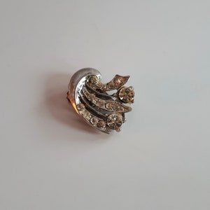 May include: A silver-toned brooch with a stylized design featuring multiple rows of clear rhinestones.