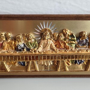 May include: A rectangular relief sculpture depicting the Last Supper. The artwork features a gold-coloured table with figures in colourful robes. The frame is a dark brown wood, and the background is gold.