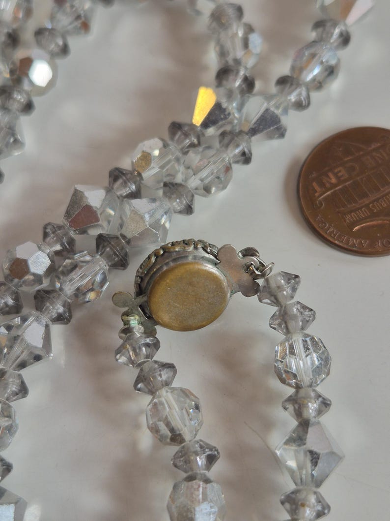 K&ouml;nnte beinhalten: Eine Halskette mit klaren, facettierten, rautenf&ouml;rmigen Glasperlen. Die Kette verf&uuml;gt &uuml;ber einen runden Metallverschluss mit messingfarbener R&uuml;ckseite und silberfarbenem Zierrand, der neben einer kupferfarbenen Ein-Cent-M&uuml;nze liegt.