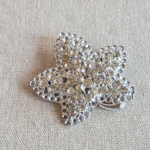 May include: A silver-toned, star-shaped brooch, encrusted with numerous clear, round rhinestones. The brooch is set against a neutral, textured background, highlighting its sparkling details. A classic accessory.