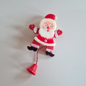 Puede incluir: Un adorno navideño vintage con forma de Papá Noel. El adorno es rojo y blanco con botas negras y una campana roja unida por una cuerda. Papá Noel tiene barba blanca, ojos azules y un gorro rojo.