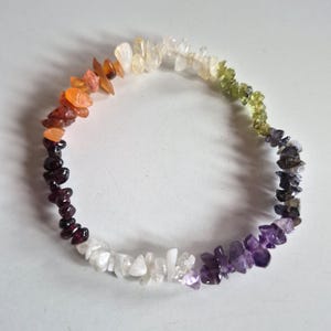 May include: A circular bracelet made of small, irregular, polished gemstone chips in a spectrum of colours including orange, red, dark purple, white, light purple, dark grey, and light green, arranged in a continuous loop on a plain surface.