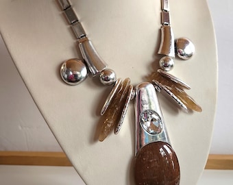 Puede incluir: Un collar de declaración plateado y marrón con un gran colgante de forma ovalada. El colgante tiene un cristal transparente en el centro y está rodeado de piedras más pequeñas, marrones, en forma de lágrima. El collar está en un maniquí blanco.
