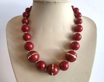 Può includere: Una collana vintage con grandi perline rotonde rosso scuro. Le perline sono separate da piccoli distanziatori color oro. Alcune delle perline più grandi hanno una fascia color oro attorno al centro. La collana è esposta su un manichino.