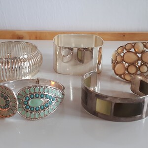 May include: Five silver bracelets with different designs. One bracelet has a geometric pattern, another has a row of white stones, another has a row of turquoise stones, and two have a smooth, plain design.