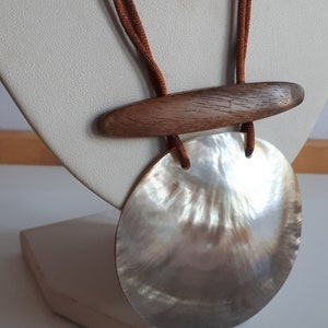 Puede incluir: Un collar de cordón marrón con un gran colgante redondo de concha blanca iridiscente y un acento de barra de madera marrón.