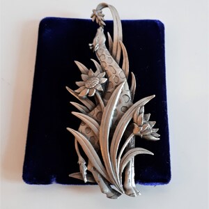 May include: A silver pewter brooch featuring a giraffe standing in a field of tall grass and flowers. The giraffe is reaching up to eat a flower.