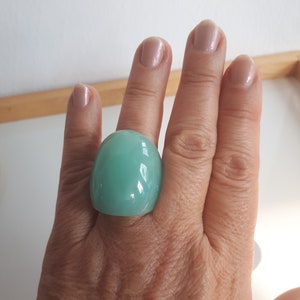 Op de afbeelding: Een grote, lichtblauwe ring met een glad, afgerond ontwerp. De ring heeft een effen kleur en lijkt gemaakt te zijn van een glanzend materiaal. De ring wordt gedragen aan een hand met gepolijste nagels.