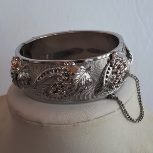 May include: Silver hinged bangle bracelet with a detailed grape and leaf design. The bracelet has an intricate engraved pattern and a safety chain. A classic piece of jewellery.