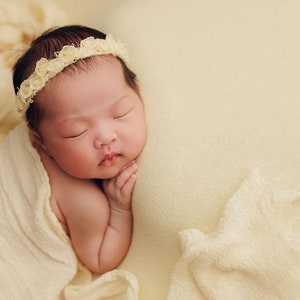 May include: A newborn baby girl sleeping peacefully on a cream-colored blanket. She is wearing a white flower headband and is swaddled in a cream-colored fabric.