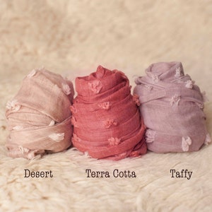 May include: Three fabric swaddles in different colors: a light pink called "Desert", a reddish pink called "Terra Cotta", and a light purple called "Taffy".