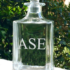 May include: A clear glass decanter with a square base and a stopper. The letters "ASE" are etched in white on the front of the decanter. The background is a blurred view of green foliage.