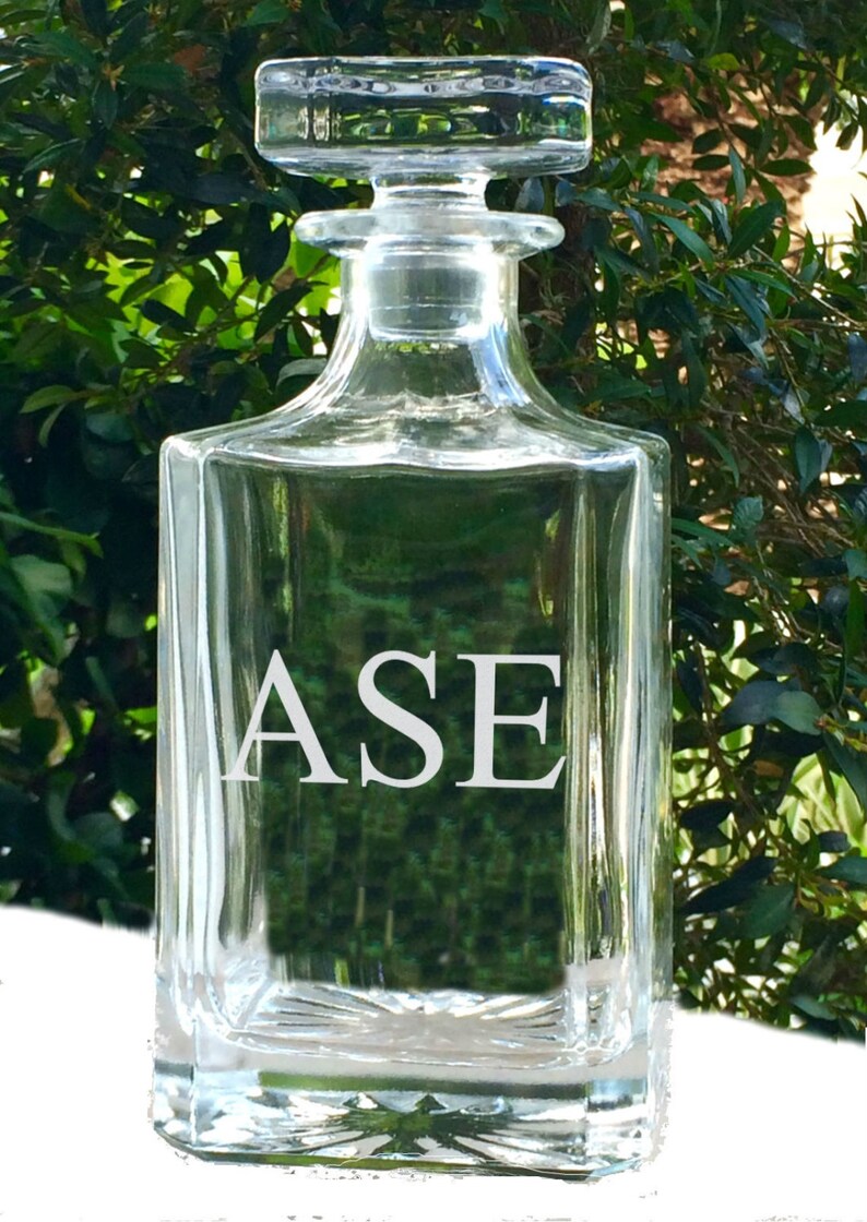 May include: A clear glass decanter with a square base and a stopper. The letters "ASE" are etched in white on the front of the decanter. The background is a blurred view of green foliage.
