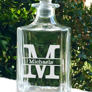 May include: A clear glass decanter with a square base and a stopper. The decanter has a white monogram "M" with the name "Michaels" below it. The background is a blurred view of green foliage.