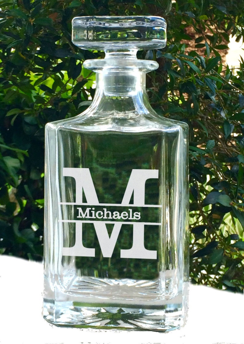 May include: A clear glass decanter with a square base and a stopper. The decanter has a white monogram "M" with the name "Michaels" below it. The background is a blurred view of green foliage.