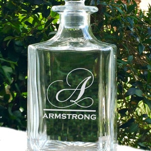 May include: Clear glass decanter with a square base and a stopper. The decanter features the name "ARMSTRONG" and the letter "A" in a decorative font. The background is a blurred view of green foliage.