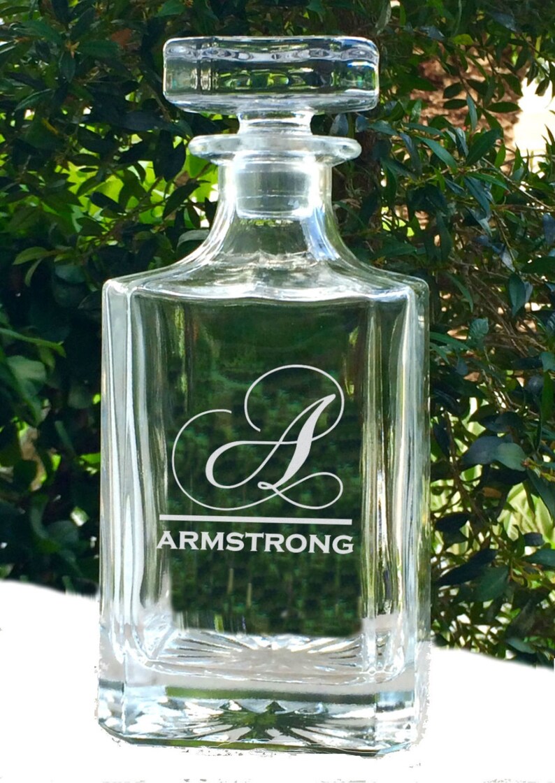 May include: Clear glass decanter with a square base and a stopper. The decanter features the name "ARMSTRONG" and the letter "A" in a decorative font. The background is a blurred view of green foliage.