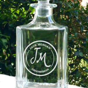 May include: A clear glass decanter with a square base and a stopper. The decanter has a circular design with the text "JACK MORGAN" above the initials "JM" and the word "GROOMSMAN" below. The background is a blurred green foliage.