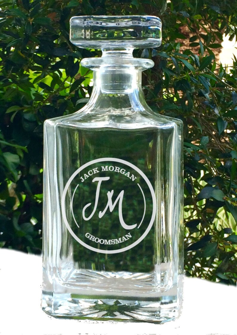 May include: A clear glass decanter with a square base and a stopper. The decanter has a circular design with the text "JACK MORGAN" above the initials "JM" and the word "GROOMSMAN" below. The background is a blurred green foliage.