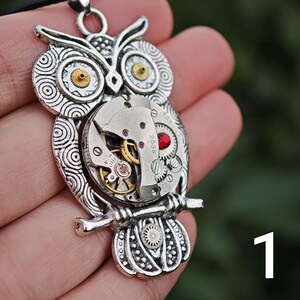 May include: Silver-toned owl pendant necklace. The owl pendant features intricate details, including watch gears and a red gem. The owl's eyes have gold-colored accents. The pendant is suspended from a black cord.