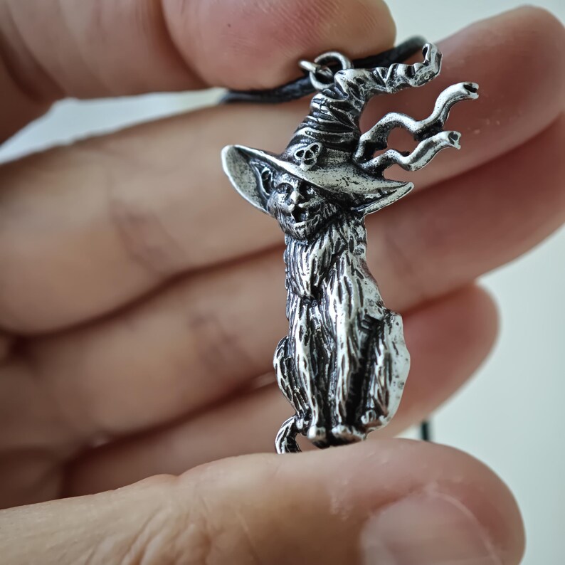 May include: A silver-colored pendant necklace featuring a cat wearing a witch's hat. The cat has a detailed, textured fur design and is seated. The witch's hat has a skull and a Celtic knot design. The pendant is suspended from a black cord.
