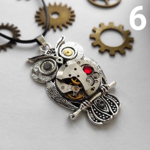May include: A silver-colored owl pendant with intricate details, featuring a watch mechanism at its center and a red gem. The pendant hangs from a black cord, with brass gears in the background. The owl is perched on a branch.