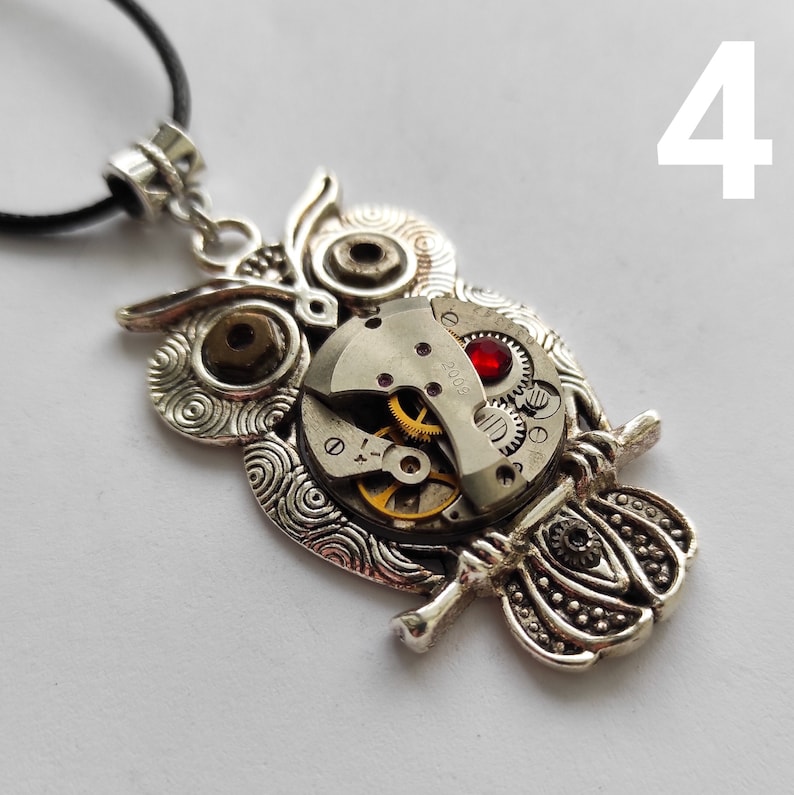May include: Silver-toned owl pendant necklace with a steampunk aesthetic. The owl's body features exposed watch gears and a red jewel. The pendant hangs from a black cord, creating a unique and artistic accessory.